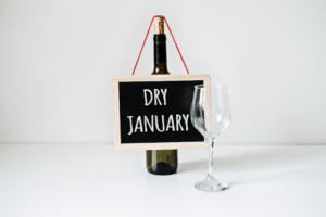 A wine bottle with a sign hanging on it that says "Dry January" with a wine glass placed in front of the bottle and sign